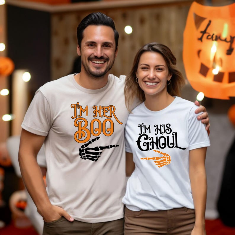 May include: A couple wearing matching Halloween t-shirts. One shirt is beige with the text "I'M HER BOO" and a skeleton hand pointing right. The other shirt is white with "I'M HIS GHOUL" and a skeleton hand pointing left.