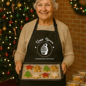 May include: A woman in a black apron with "Claire Brown Handmade Pastries" holds a tray of festive cookies. The tray includes gingerbread men, stars, and Christmas trees. A Christmas tree and wreath are visible in the background.