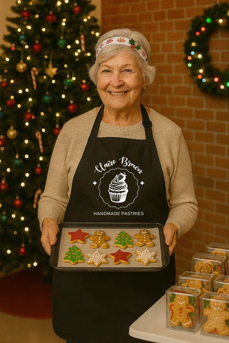 May include: A woman in a black apron with "Claire Brown Handmade Pastries" holds a tray of festive cookies. The tray includes gingerbread men, stars, and Christmas trees. A Christmas tree and wreath are visible in the background.