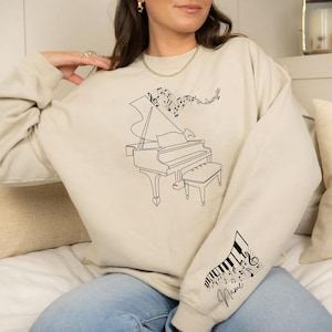 May include: Beige sweatshirt featuring a black line drawing of a grand piano with musical notes. The design includes a piano bench and musical notes floating above the piano. The sleeve has a piano keyboard and musical notes with the word "Name" in cursive.