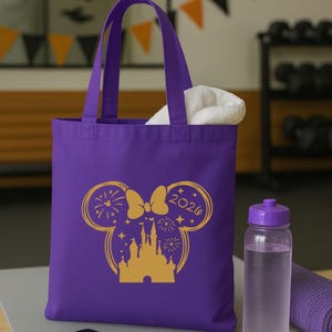 Puede incluir: Un bolso tote morado con un gr&aacute;fico dorado de un castillo, fuegos artificiales y un lazo con forma de orejas de Mickey Mouse. El texto "2020" est&aacute; en la oreja derecha. Una toalla blanca est&aacute; dentro de la bolsa. Una botella de agua morada y una esterilla de yoga enrollada.