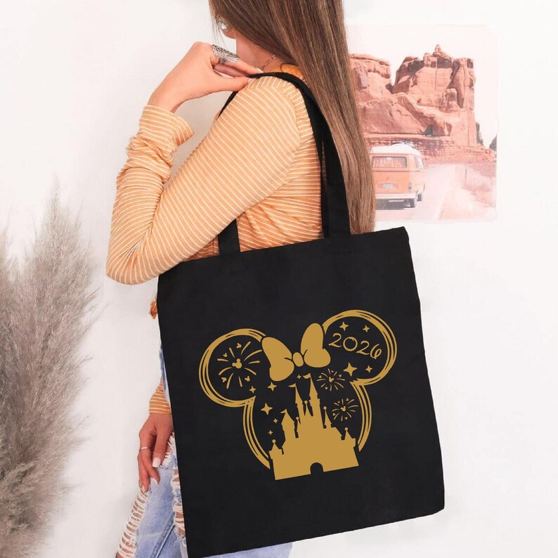 Puede incluir: Bolso tote negro con un dise&ntilde;o dorado de Minnie Mouse, que incluye una silueta de castillo, fuegos artificiales y el a&ntilde;o 2026. El bolso tiene forma rectangular y dos asas, perfecto para llevar art&iacute;culos.