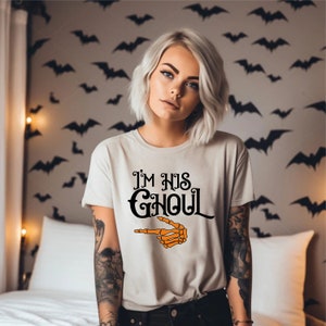 May include: A light beige t-shirt with the text "I'M HIS GHOUL" in black script and a pointing orange skeleton hand. The shirt is a Halloween-themed design.