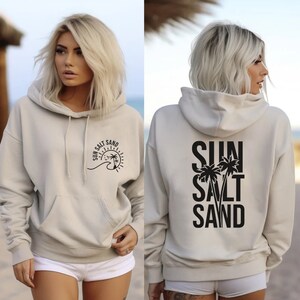 May include: A light beige hoodie with a black graphic on the front that says "Sun Salt Sand" with a sun, wave, and palm trees. The back of the hoodie has a black graphic that says "Sun Salt Sand" with palm trees.