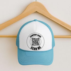 May include: A blue and white trucker hat with a curved brim. The front panel is white with a circular design that includes a QR code and the text "COMPANY NAME" and "SCAN ME" in black. The hat is hanging on a wooden hanger.