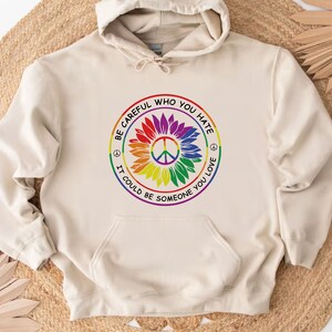 Pu&ograve; includere: Una felpa con cappuccio beige con una grafica di girasole arcobaleno e un simbolo di pace. La grafica include il testo "Fai attenzione a chi odi, potrebbe essere qualcuno che ami".