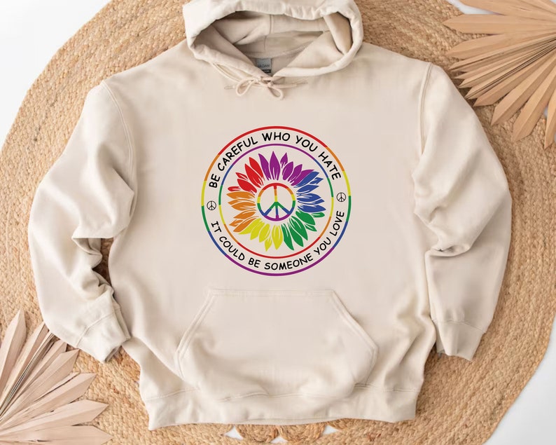 Pu&ograve; includere: Una felpa con cappuccio beige con una grafica di girasole arcobaleno e un simbolo di pace. La grafica include il testo "Fai attenzione a chi odi, potrebbe essere qualcuno che ami".