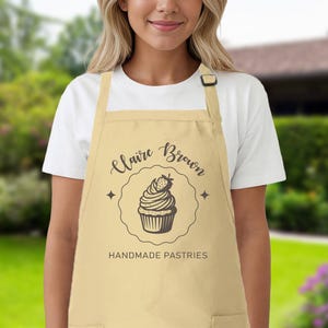 May include: A beige apron featuring the name "Claire Brown" and the words "Handmade Pastries" in black script. A cupcake illustration with a strawberry is in the centre. The apron has a pocket and an adjustable neck strap.