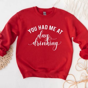 K&ouml;nnte beinhalten: Ein rotes Sweatshirt mit dem wei&szlig;en Text "YOU HAD ME AT day drinking" in einer Schreibschrift. Das lang&auml;rmlige Oberteil ist aus einem weichen Material gefertigt und eignet sich f&uuml;r Freizeitkleidung. Das Sweatshirt hat eine einfarbige rote Farbe.