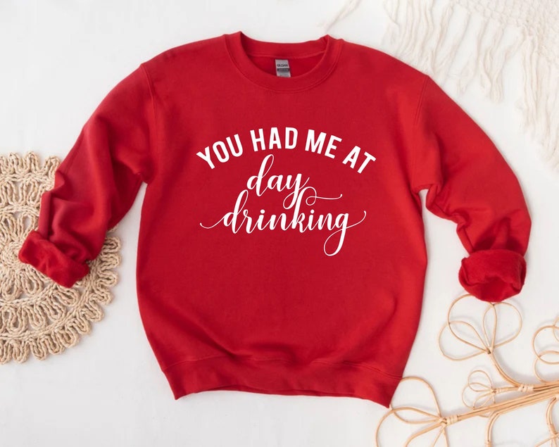K&ouml;nnte beinhalten: Ein rotes Sweatshirt mit dem wei&szlig;en Text "YOU HAD ME AT day drinking" in einer Schreibschrift. Das lang&auml;rmlige Oberteil ist aus einem weichen Material gefertigt und eignet sich f&uuml;r Freizeitkleidung. Das Sweatshirt hat eine einfarbige rote Farbe.