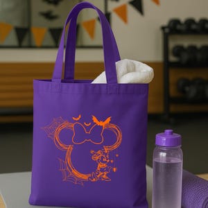 Puede incluir: Un bolso tote morado con un gr&aacute;fico naranja de la silueta de un rat&oacute;n con un lazo, telara&ntilde;as, murci&eacute;lagos y un peque&ntilde;o personaje de rat&oacute;n. El bolso tiene dos asas y contiene una toalla blanca.