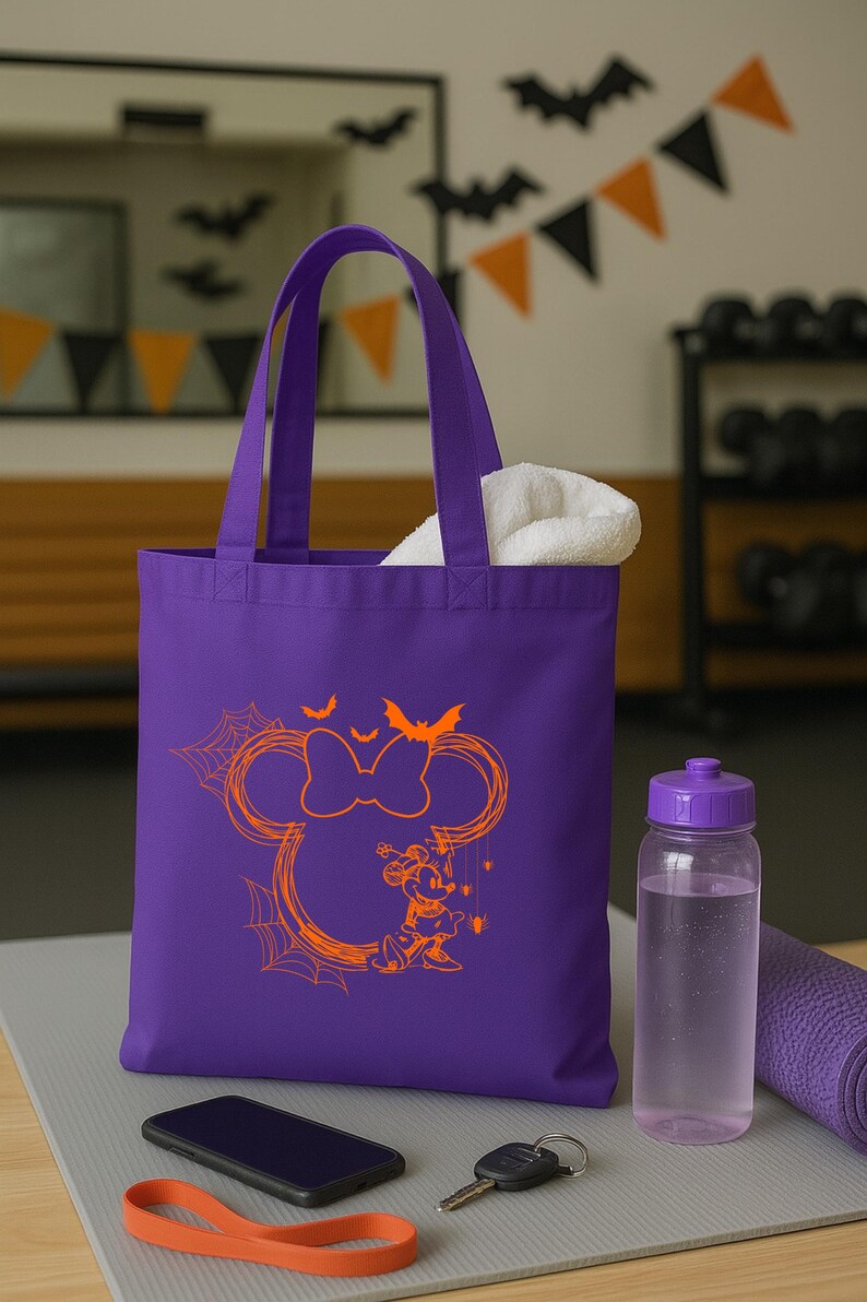 Puede incluir: Un bolso tote morado con un gr&aacute;fico naranja de la silueta de un rat&oacute;n con un lazo, telara&ntilde;as, murci&eacute;lagos y un peque&ntilde;o personaje de rat&oacute;n. El bolso tiene dos asas y contiene una toalla blanca.