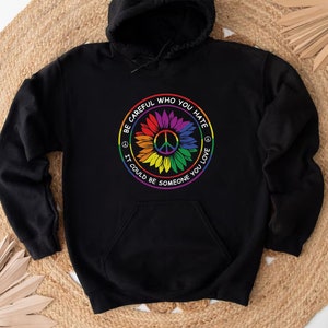Pu&ograve; includere: Felpa con cappuccio nera con un simbolo di pace arcobaleno e un design a girasole. Il testo recita "Fai attenzione a chi odi, potrebbe essere qualcuno che ami."
