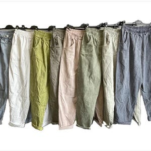 May include: A collection of nine pairs of casual trousers hanging on black hangers, featuring an elasticated waistband, side pockets, and a relaxed fit. The trousers are available in various solid colours including light blue, white, olive, grey, pink, and sage.