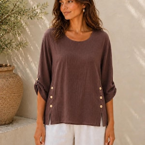 May include: A dark brown long-sleeve blouse featuring a round neckline, three-quarter length sleeves with button-tab cuffs, and side slits accented with three small light-colored buttons on each side, paired with loose-fitting white trousers.