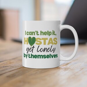 May include: White ceramic mug with a green and gold design. The mug has the text "I can't help it, HOSTAS get lonely by themselves".