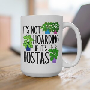 May include: White ceramic mug with the text "IT'S NOT HOARDING IF IT'S HOSTAS" and illustrations of green plants in blue, pink, and purple pots. The mug has a large handle and is on a wooden surface.