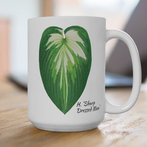 May include: White ceramic mug featuring a detailed illustration of a green and white leaf. The leaf design is centered on the mug, with the text "H. 'Sharp Dressed Man'" printed below the leaf. The mug has a curved handle.