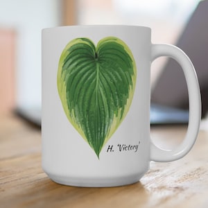 May include: White ceramic mug with a green and yellow hosta leaf illustration in the shape of a heart. The text 'H. 'Victory'' is printed below the leaf.