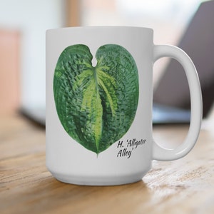 May include: White ceramic mug with a green and yellow leaf design. The leaf is shaped like a heart and has a textured surface. The text "H. 'Alligator Alley'" is printed below the leaf.