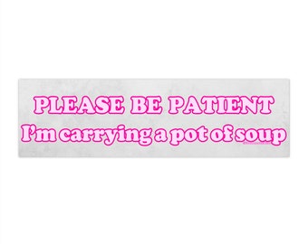 May include: A rectangular bumper sticker with the text "PLEASE BE PATIENT I'm carrying a pot of soup" in bright pink letters. The background is a light gray, textured pattern.