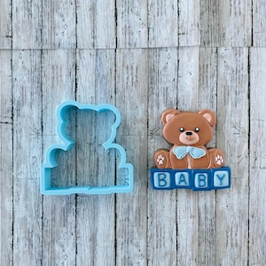 May include: A blue plastic cookie cutter shaped like a teddy bear sitting on top of a block that says "BABY".  The teddy bear is holding a blue bow tie.  The cookie cutter is next to a decorated cookie of a teddy bear sitting on top of a block that says "BABY".