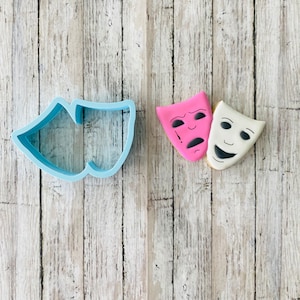 May include: A blue plastic cookie cutter shaped like two theater masks. Next to it are two decorated sugar cookies, one pink with a sad face and one white with a happy face.