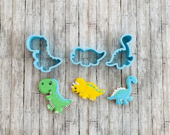 May include: A collection of dinosaur-themed cookie cutters and decorated cookies. The cutters are light blue plastic, and the cookies are decorated in green, yellow, and blue icing. The cookies feature a T-Rex, a Triceratops, and a Brontosaurus.