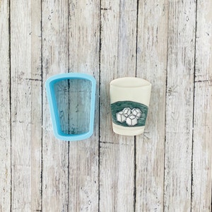 May include: A blue plastic cookie cutter shaped like a glass with ice cubes. Next to it is a decorated cookie that looks like a glass of iced tea.