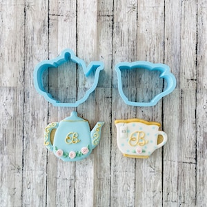 May include: Two blue cookie cutters, one shaped like a teapot and the other like a teacup, are shown next to two decorated cookies, one shaped like a teapot and the other like a teacup. The cookies are decorated with blue and white icing and gold accents.