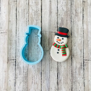 May include: A blue plastic snowman cookie cutter next to a decorated gingerbread snowman cookie with a black top hat, green and red scarf, and black eyes and buttons.