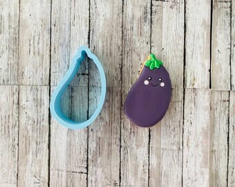 May include: A blue plastic eggplant cookie cutter and a purple and green decorated sugar cookie shaped like an eggplant with a smiling face.