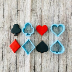 May include: A set of eight cookie cutters in the shape of playing card suits. The cutters are made of blue plastic and include a heart, diamond, spade, club, and three solid shapes: a red heart, a red diamond, and a black spade.