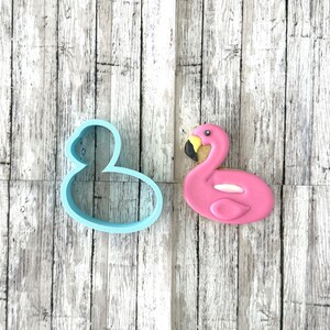 Puede incluir: Una galleta rosa con forma de flamenco con pico negro y amarillo y detalles blancos. Junto a ella, un cortador de galletas azul con forma de flamenco. La galleta está sobre un fondo de madera.