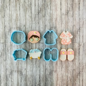 May include: A set of eight blue cookie cutters in various shapes, including a bath tub, slippers, a robe, and a person with a face mask. The cookie cutters are arranged on a light brown wooden surface.