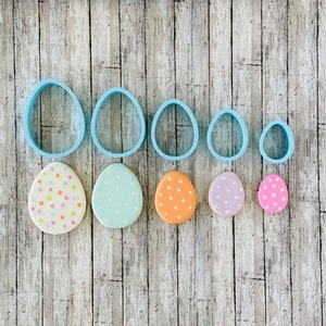Könnte beinhalten: Ein Set mit sechs blauen Plastik-Oster-Eier-Ausstechern und sechs dekorierten Ostereiern. Die Eier sind mit Pastellfarben und weißen Punkten verziert.