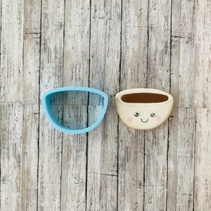 May include: A blue plastic cookie cutter shaped like a bowl, and a white cookie decorated to look like a bowl with a brown liquid inside and a smiling face.