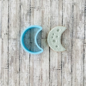 Puede incluir: Dos formas de media luna, una es un cortador de galletas de plástico azul claro y la otra es una luna de cerámica gris texturizada con cráteres.