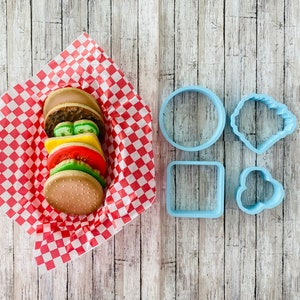 Puede incluir: Una pila de galletas de azúcar decoradas con forma de hamburguesa sobre papel de cuadros rojos y blancos. A la derecha de las galletas se muestran cuatro cortadores de galletas de plástico azul: un círculo, un cuadrado, una nube y un corazón.