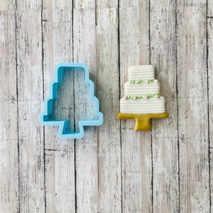 Puede incluir: Un cortador de galletas de plástico azul con forma de pastel de boda, junto a una galleta de pastel de boda decorada con glaseado blanco y flores rosas.