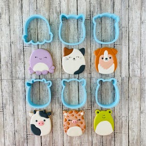 May include: A collection of blue plastic cookie cutters in various animal shapes, including an octopus, cat, dog, cow, giraffe, and frog. The cut-out cookies are decorated with colorful icing, showcasing the animal designs. The cutters are arranged on a light wood-grain surface.