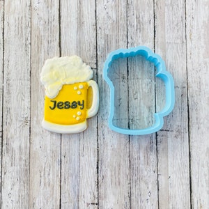 May include: A blue plastic cookie cutter shaped like a beer mug with a white foam top. Next to it is a decorated sugar cookie in the shape of a beer mug with the name "Jessy" written on it.