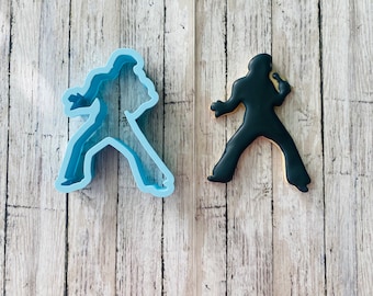May include: Two cookie cutters, one blue and one black, shaped like a silhouette of Elvis Presley. The blue cutter is on the left and the black cutter is on the right. Both cutters are on a wooden background.