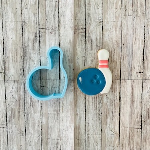 May include: A blue plastic cookie cutter shaped like a bowling pin and a decorated sugar cookie shaped like a bowling pin and ball.