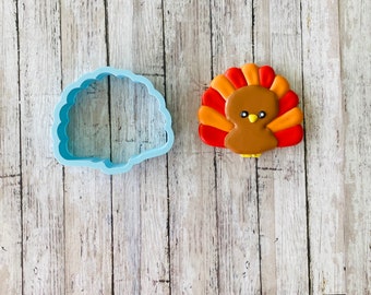 May include: A blue plastic cookie cutter shaped like a turkey, next to a decorated sugar cookie shaped like a turkey with brown, orange, and red icing.