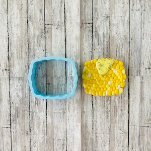 May include: A blue plastic cookie cutter shaped like a rectangle with rounded corners. Next to it is a yellow and white cookie decorated with a yellow and grey piece of fondant.