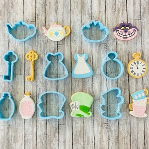 May include: Assorted cookie cutters in the theme of Alice in Wonderland. Includes a teapot, dress, key, Cheshire Cat, pocket watch, and Mad Hatter's hat. The cutters are blue, yellow, pink, and white.