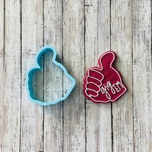 May include: A red cookie cutter in the shape of a thumbs up hand with the text "gig 'em" written on it.  A blue plastic cookie cutter in the same shape is next to it.