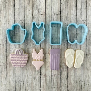 May include: A set of six cookie cutters in the shape of a beach bag, swimsuit, towel, flip flops, and a beach bag with a striped pattern. The cutters are light blue and the cookies are decorated with purple, white, and pink icing.