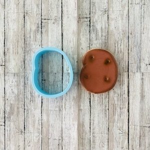 May include: A blue plastic cookie cutter shaped like a heart and a brown cookie decorated with four white dots.